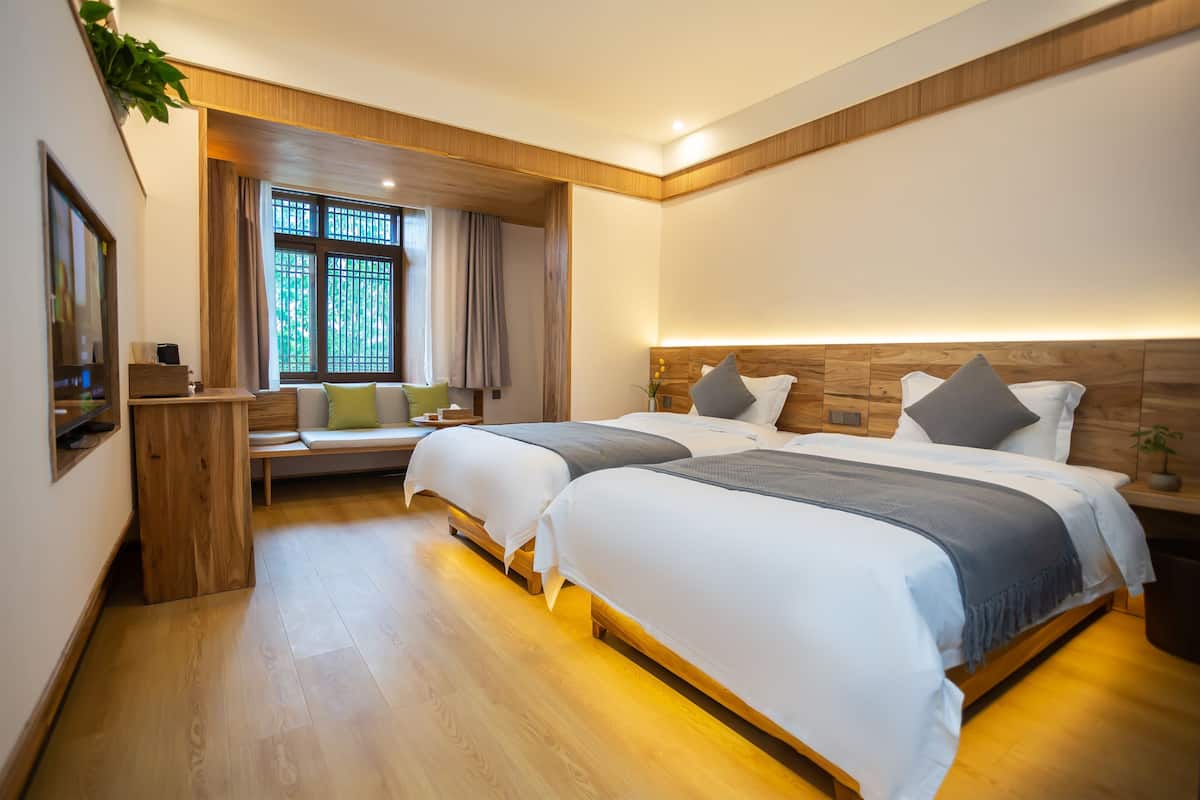Una habitación de hotel moderna con dos camas, cabecera de madera, una pequeña zona de estar y un ventanal con cortinas.
