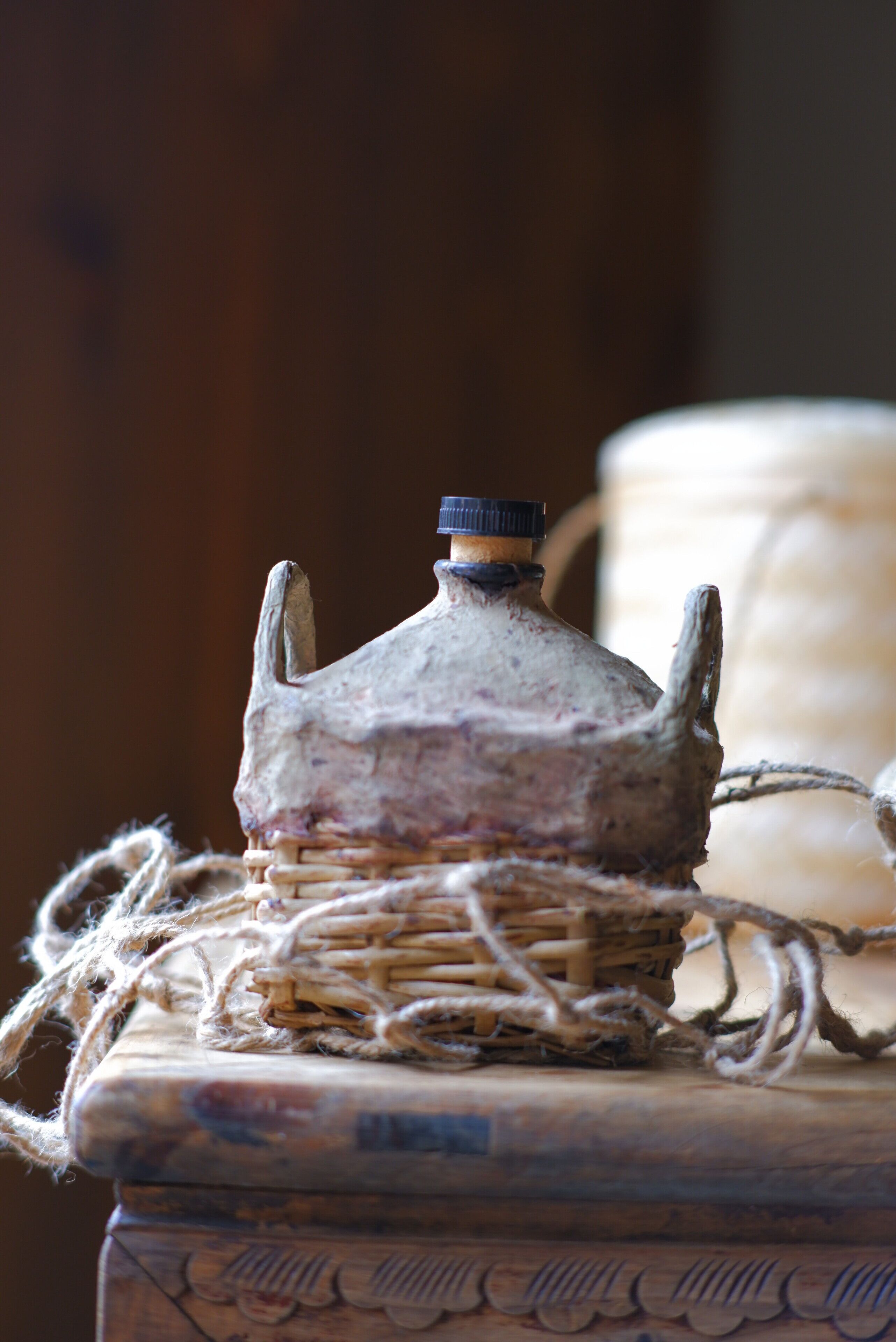 A rustic, old-fashioned bottle with a metal cap, tied with a rope, placed on a wooden surface with carved details.