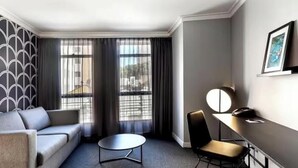 A hotel room with a sofa, a round table, a chair, a desk, and a large window with curtains. - KozyGuru | Haymarket | Best Location Studio (Haymarket)