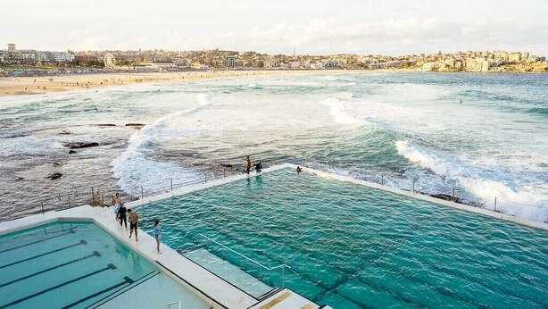 A coastal swimming area with a pool adjacent to the sea, a beach in the background, and a town across the water.