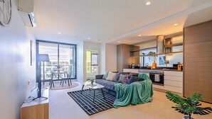 A modern apartment with a dining area, a sofa, a kitchen, and a large window. - KozyGuru | Abbotsford | Cosy 1Bed Apt (Abbotsford)