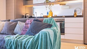 A modern living room with a sofa covered in a teal blanket, a kitchen area with appliances, and a hanging plant. - KozyGuru | Abbotsford | Cosy 1Bed Apt (Abbotsford)