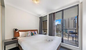 A hotel room with a large bed, a nightstand, a desk, a chair, and a balcony with a city view.