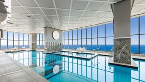 An indoor swimming pool with clear blue water, large windows offering ocean views, and marble accents on the pool's edge.