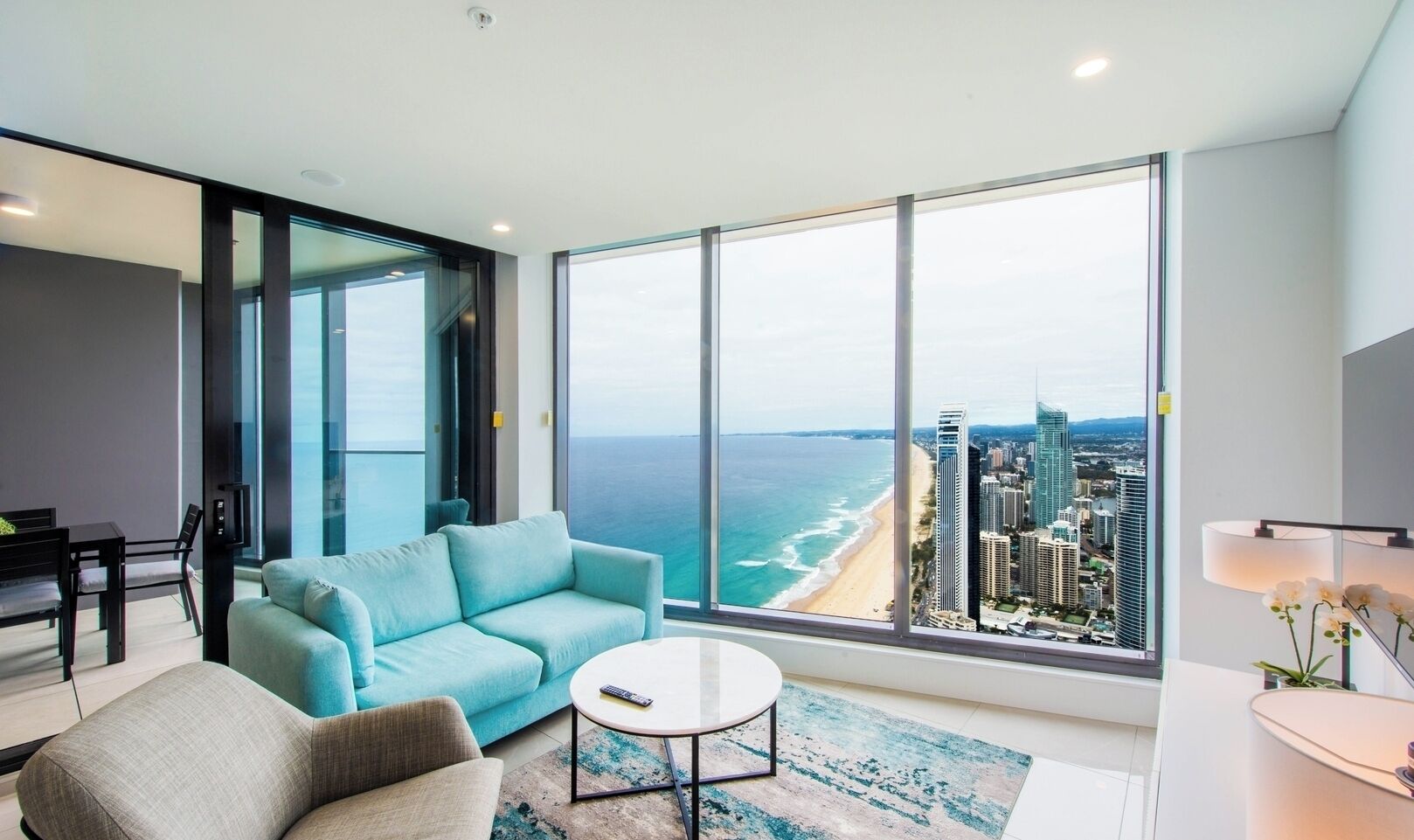 A modern living room with a large window, a sofa, a coffee table, and a view of the ocean and city skyline.