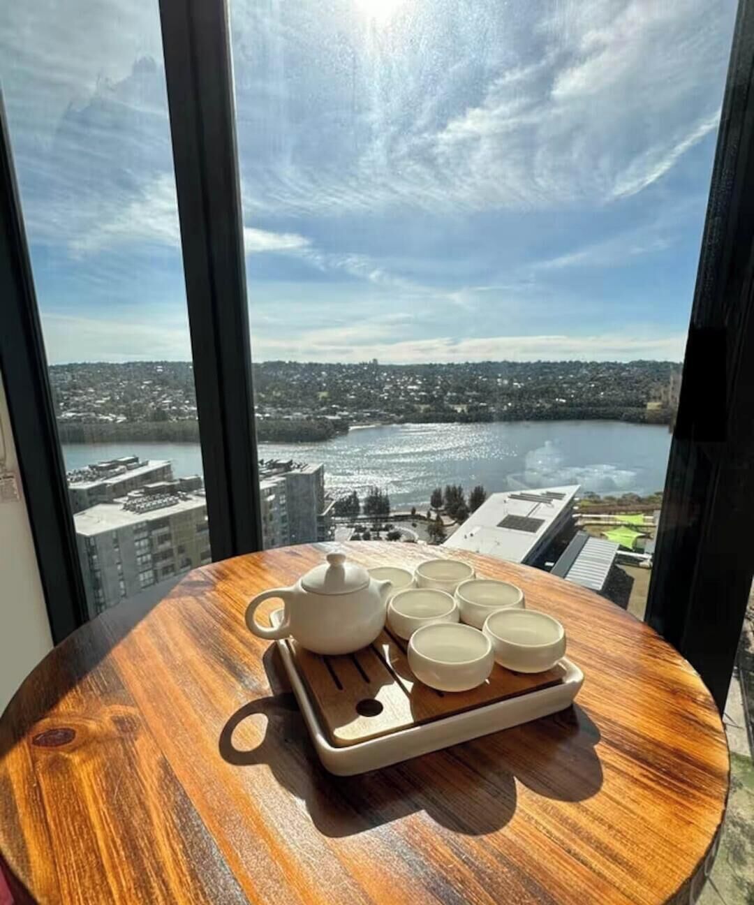 Une table ronde en bois, sur laquelle se trouve un plateau avec une théière et six tasses, est placée près d’une fenêtre donnant sur un paysage urbain et une étendue d’eau.