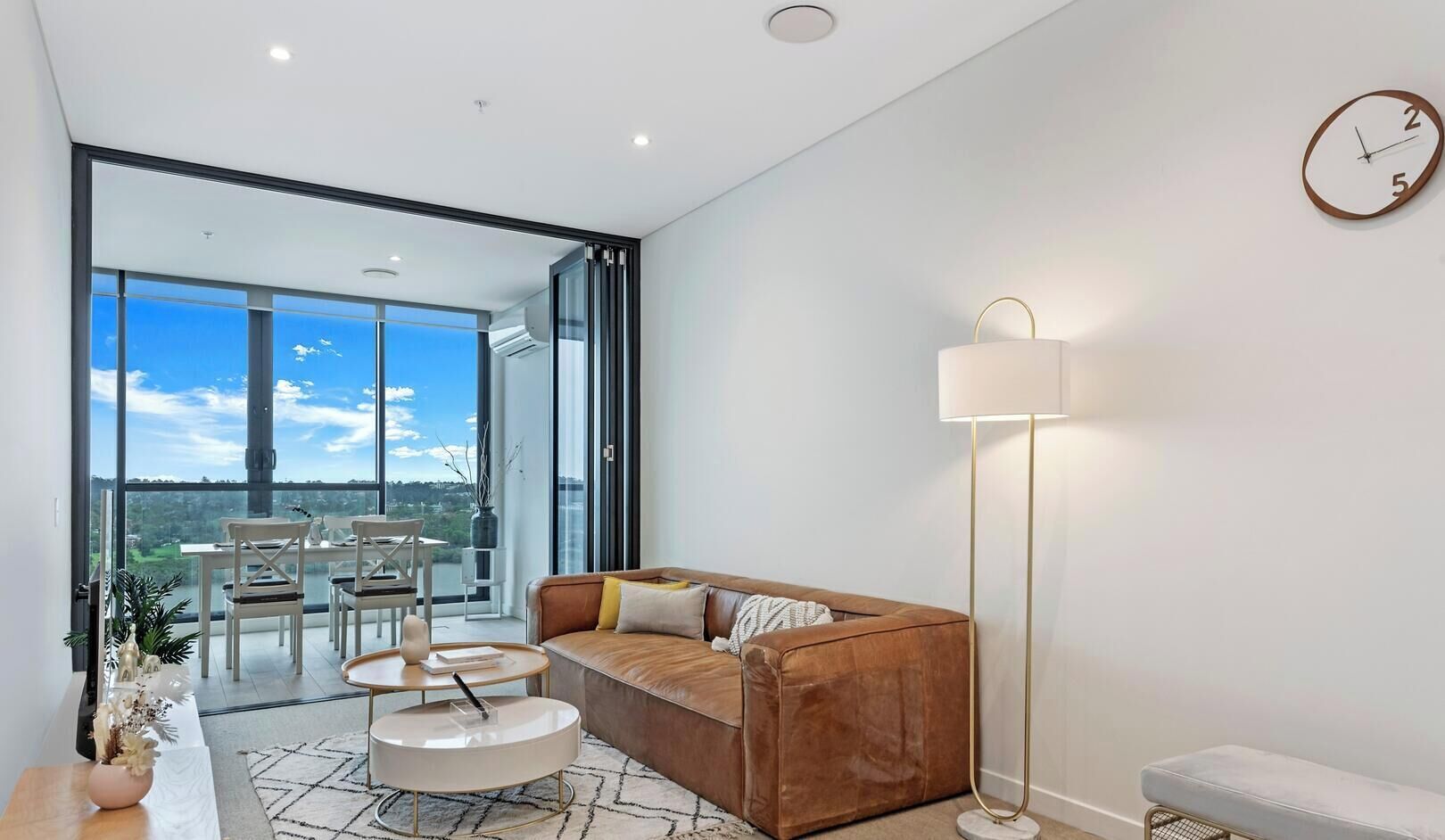 A spacious living room with a leather sofa, a round coffee table, and a floor lamp. A dining area with a table and chairs is visible through large windows.