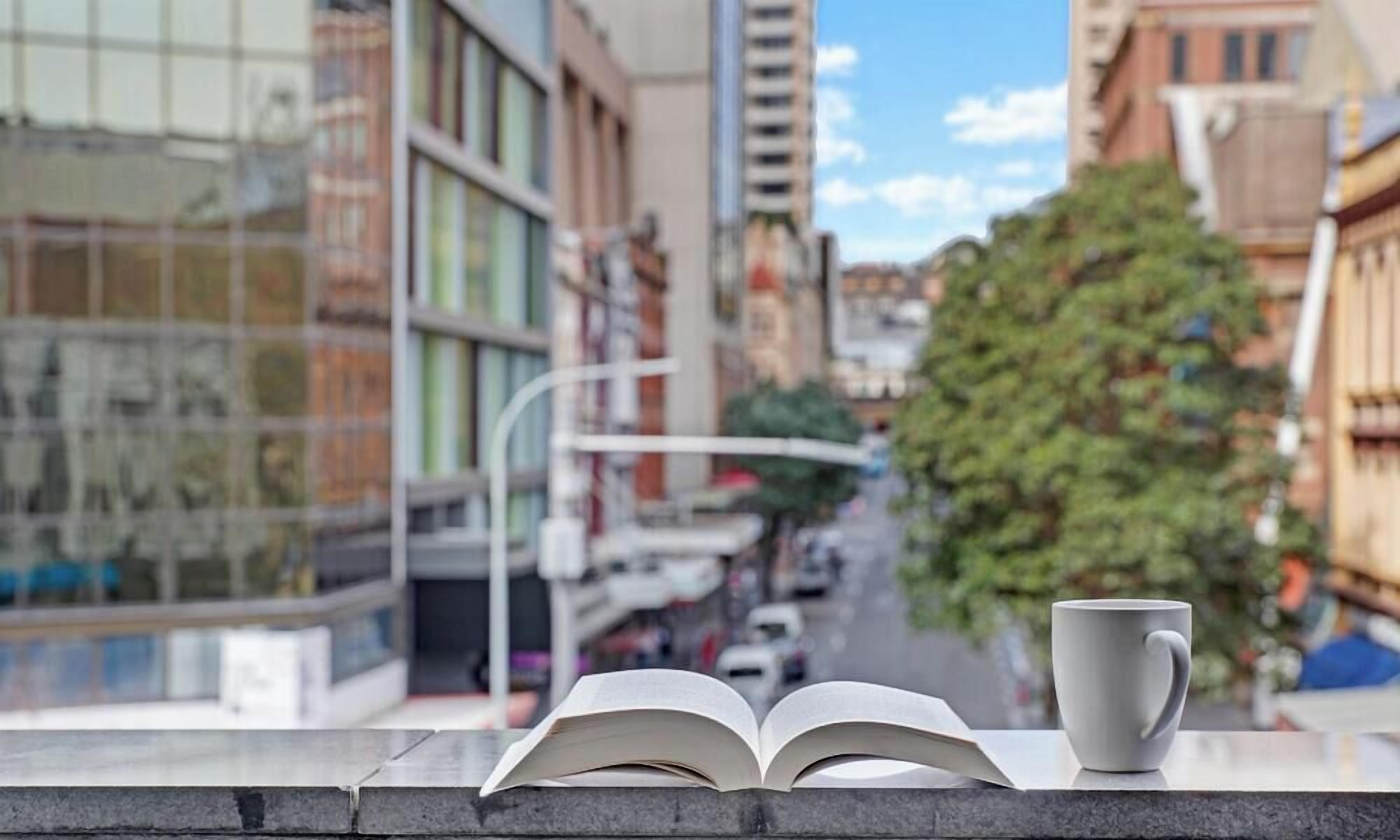 An open book and a mug on a ledge with a city street view in the background.