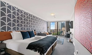 A hotel room with a large bed, a sofa, a desk, and a balcony with a city view.