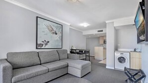 Una sala de estar moderna con un sofá seccional gris, un comedor con mesa y sillas, una cocina con lavadora y un televisor montado.