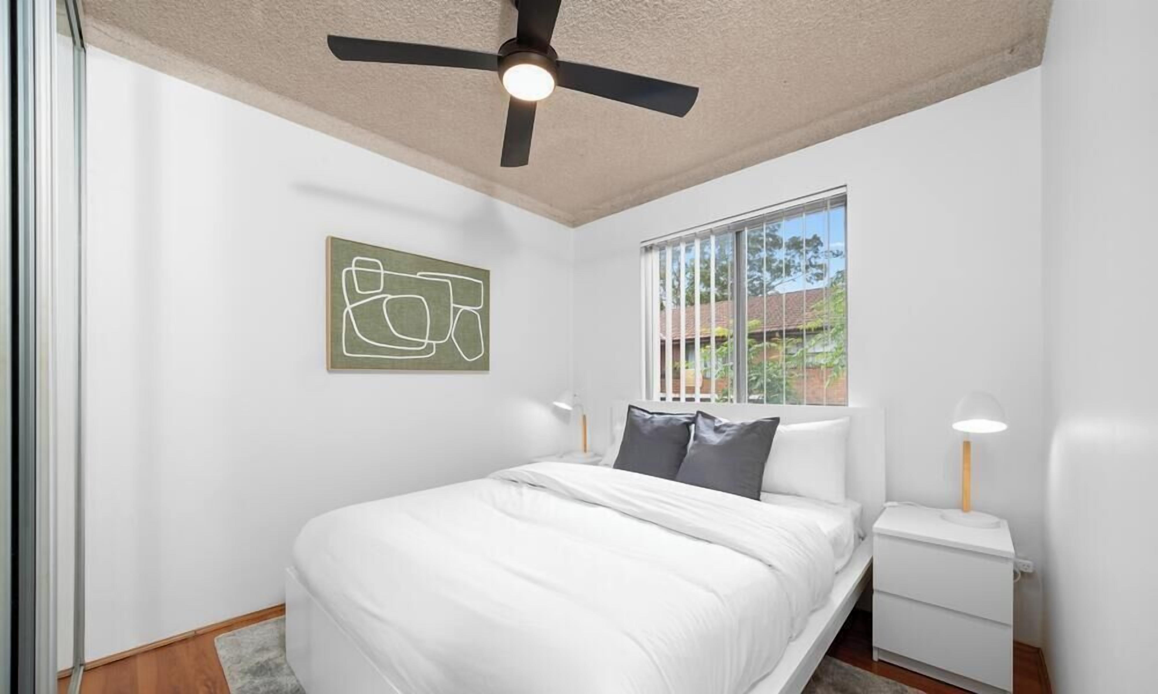 A bedroom with a bed, a ceiling fan, a window with blinds, a bedside table, and a wall-mounted artwork.