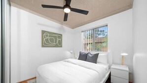 A bedroom with a bed, a ceiling fan, a window with blinds, a bedside table, and a wall-mounted artwork. - KozyGuru | Westmead  | 2Bed Unit + Free Parking (Westmead)