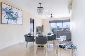Un espace de salle à manger moderne avec une table en verre, quatre chaises, un lustre et une vue sur la ville grâce à de grandes fenêtres.