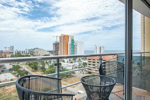A balcony with two wicker chairs, a view of a cityscape with buildings and a distant shoreline.