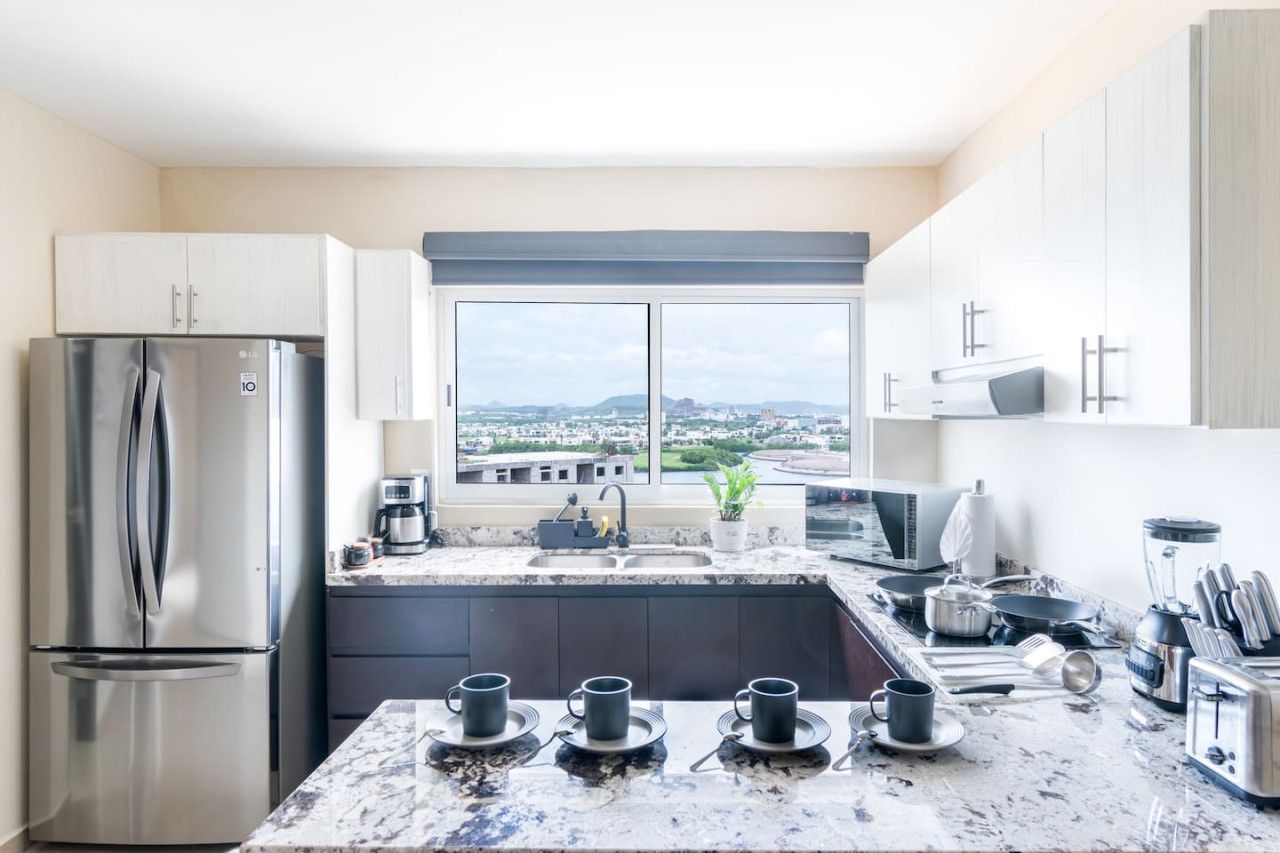 A modern kitchen with a stainless steel refrigerator, a large window with a city view, and a marble countertop.