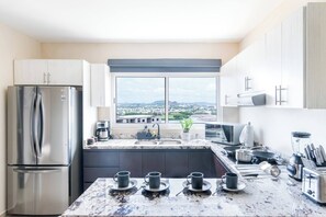A modern kitchen with a stainless steel refrigerator, a large window with a city view, a sink, and a countertop with various kitchen items.