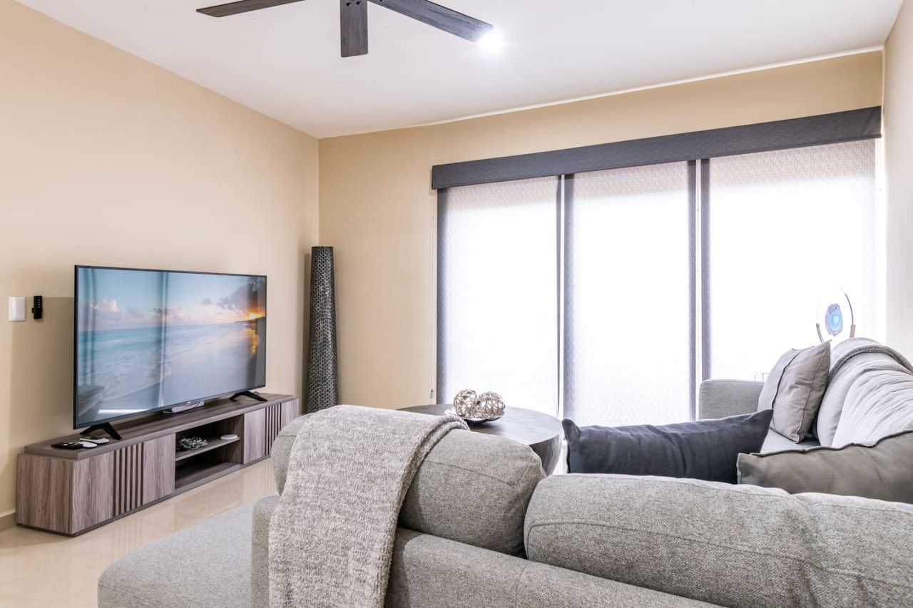 A modern living room with a flat-screen TV on a wooden stand, a grey sofa, and a large window with curtains.