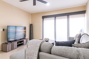 Una sala de estar moderna con un televisor de pantalla plana sobre un soporte de madera, un sofá gris y un ventanal con cortinas.