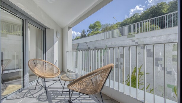 A balcony with two wicker chairs and a small table, overlooking a street with a white fence and greenery.
