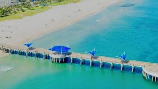 Luchtbeeld van een kustgebied met een steiger die het water in reikt, met daaraan meerdere blauwe parasols.