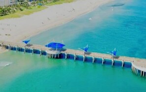 An aerial view of a coastal area with a pier extending into the water, featuring several blue umbrellas.