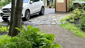 Une voiture blanche est garée dans une allée menant à une maison entourée d’une végétation luxuriante, avec un chemin de pierres qui y conduit.