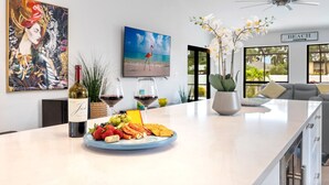 Une cuisine moderne avec un coin repas, comprenant un grand îlot central agrémenté d’un plateau de fruits, de verres à vin et d’une bouteille de vin. On y trouve également des plantes décoratives, un tableau et une vue sur la plage grâce à de grandes fenêtres.