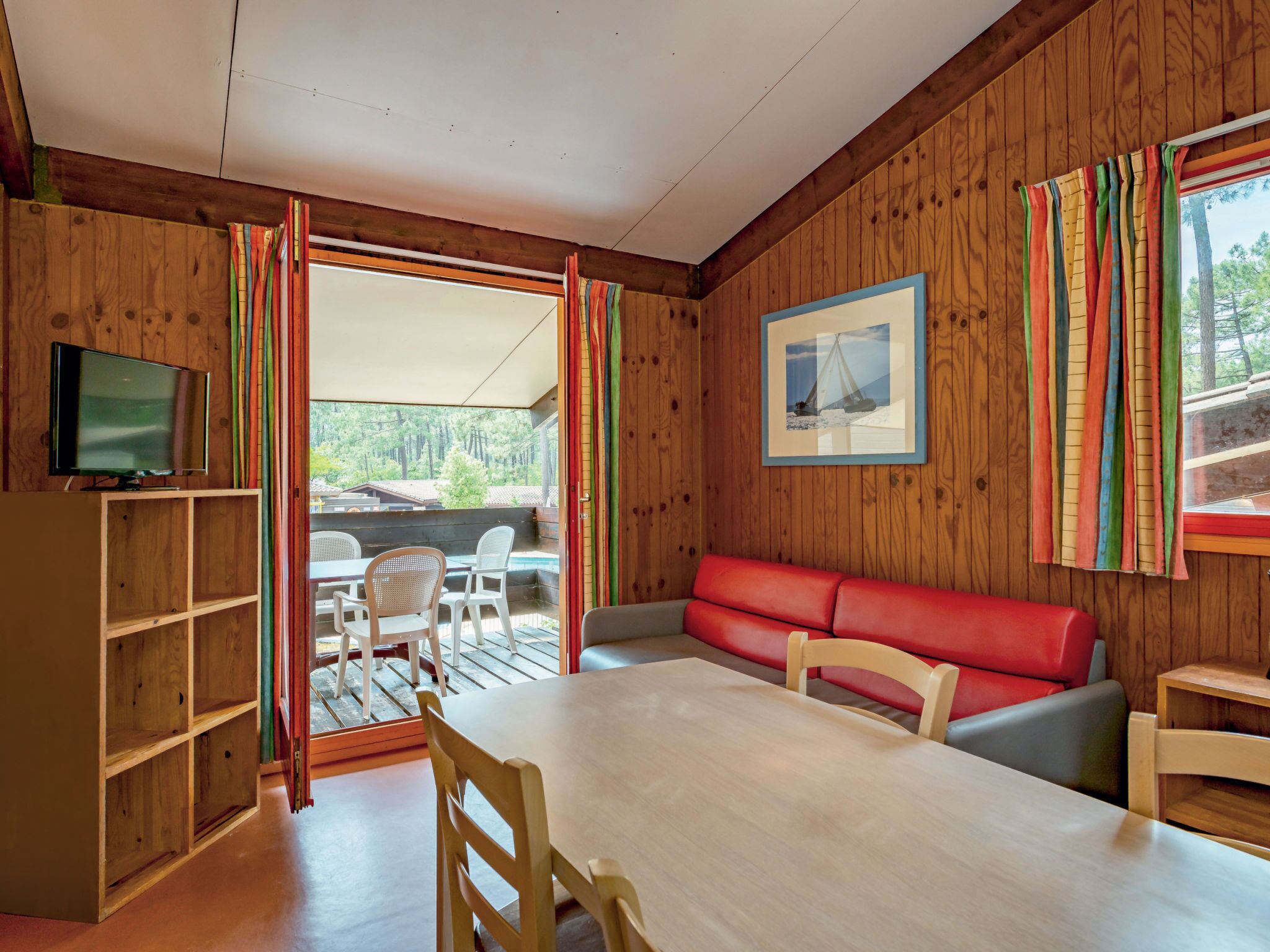 Een kamer met houten lambrisering, een eettafel, stoelen, een rode bank, een televisie op een houten kast, en een raam met kleurrijke gordijnen.