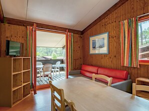 A wooden-paneled room with a dining table, chairs, a red sofa, a TV on a wooden cabinet, and a window with colorful curtains.