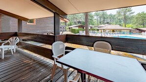 A covered outdoor seating area with wooden benches and white plastic chairs, overlooking a swimming pool and a building with a tiled roof. - Les Sentes by Interhome (Maubuisson)