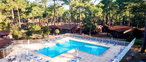 Zona de piscina con sillas de descanso y mesas, rodeada de árboles y edificios.