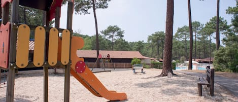 Un parque infantil con tobogán y estructuras para escalar, rodeado de árboles y una zona de arena.