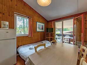 A cozy cabin-style room with a wooden interior, a bed, a dining table, chairs, a TV, and a window with curtains. - Les Sentes by Interhome (Maubuisson)