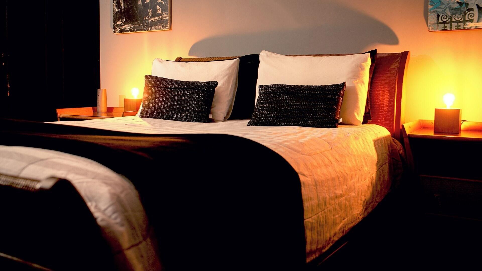 Uma cama com edredom branco, duas almofadas pretas e cabeceira de madeira. Há duas mesas de cabeceira com velas.