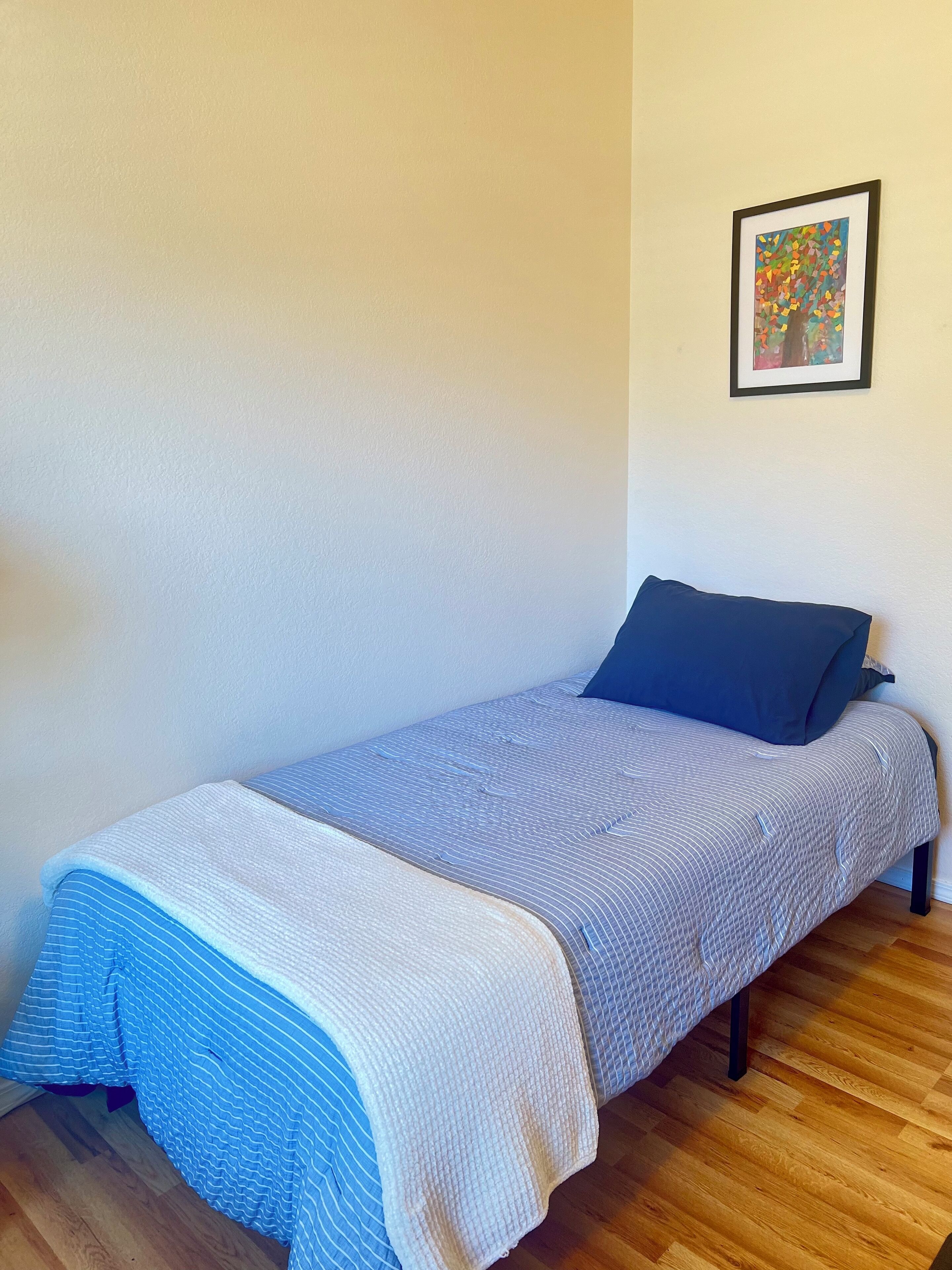 Uma cama de solteiro com colcha listrada em azul e branco e um cobertor branco, travesseiro azul escuro, e uma pintura abstrata emoldurada na parede.