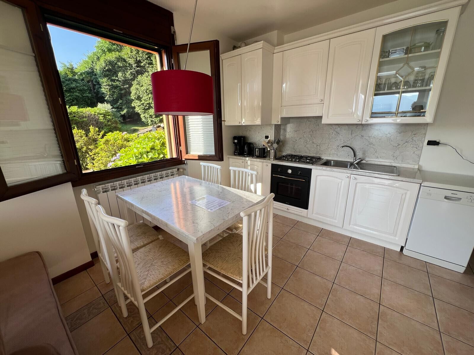 A kitchen with white cabinets, a dining table with chairs, a red hanging light, and a view of greenery outside.