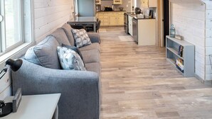 Amplia sala de estar con una escalera de madera que sube a una entreplanta, un sofá gris y una cocina al fondo.