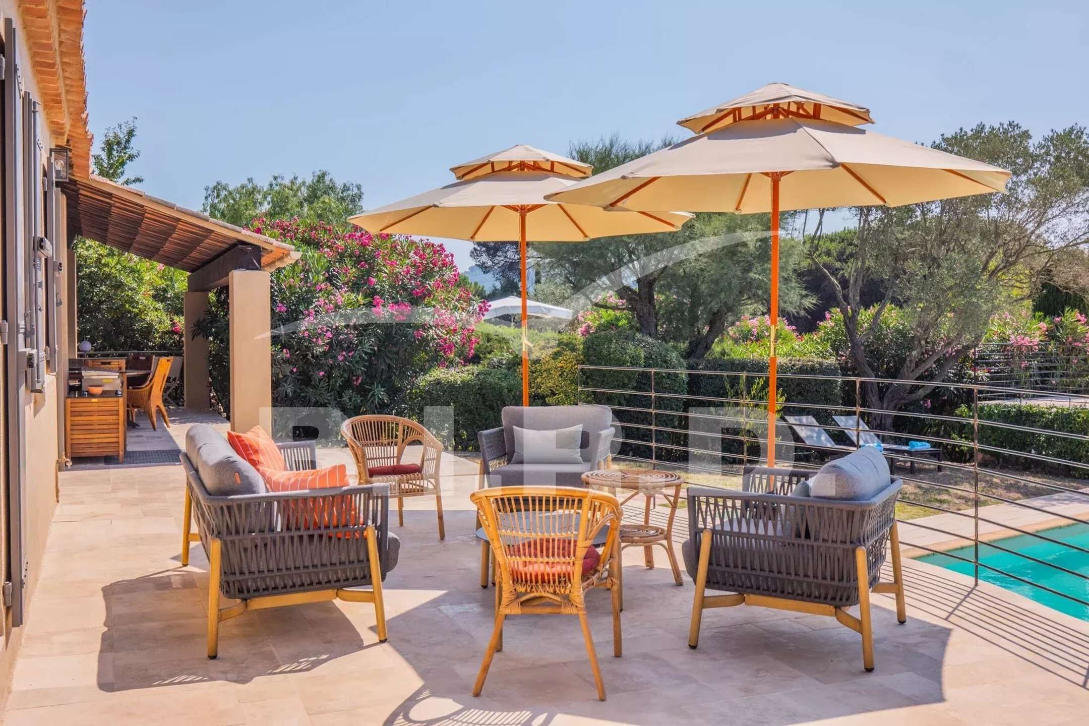 An outdoor patio with wicker furniture and large umbrellas, a pool, and lush greenery.