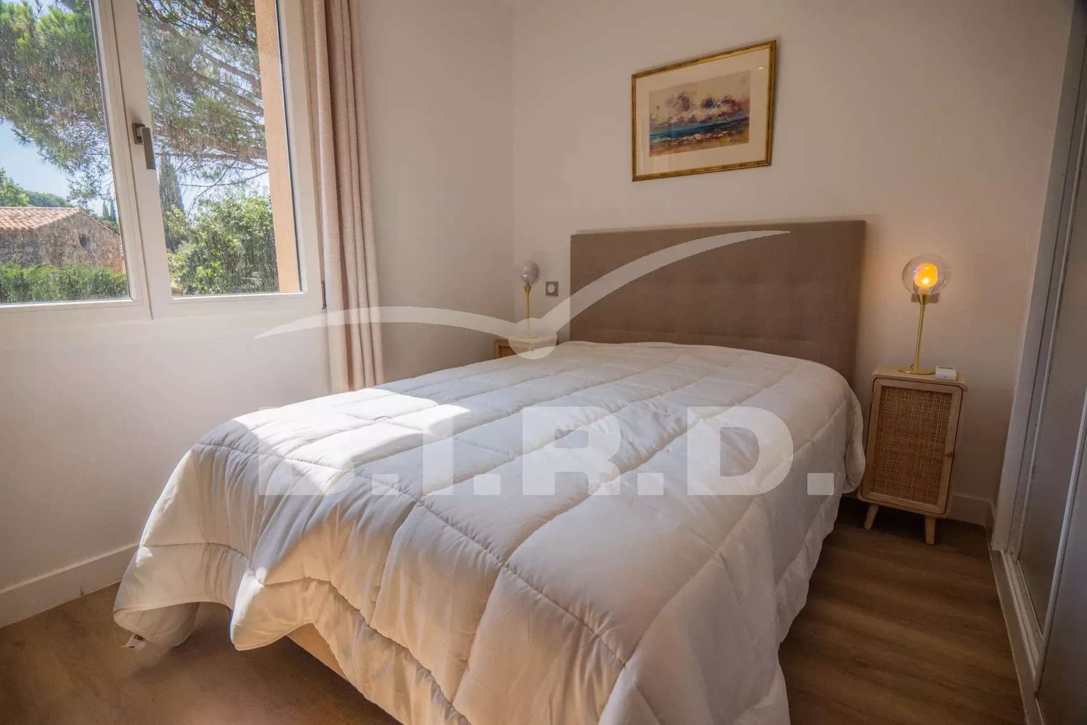 A bedroom with a large bed, a bedside table with a lamp, a framed picture on the wall, and a window with curtains.