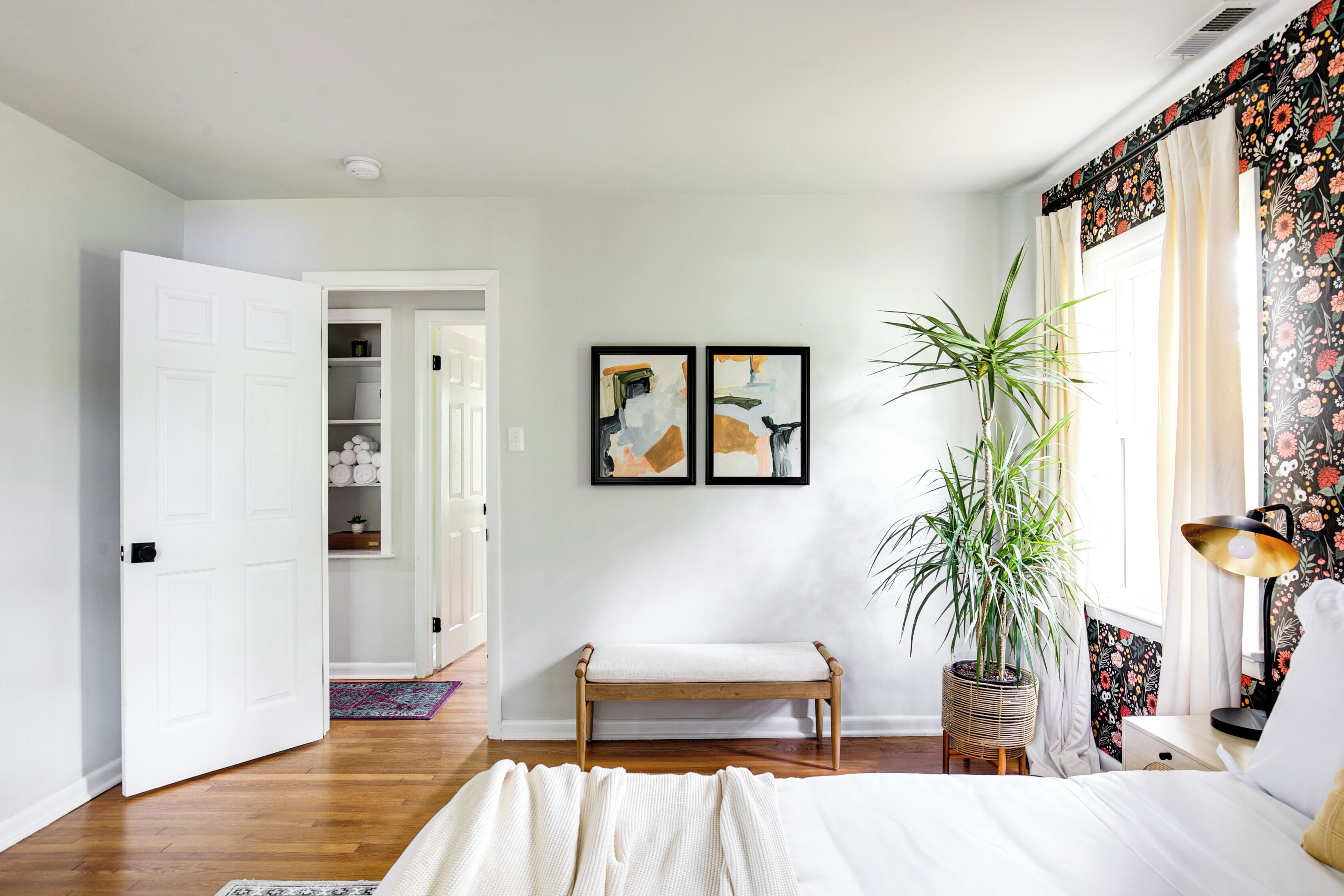 A bedroom with a bed, a bench, a potted plant, and two framed artworks on the wall.