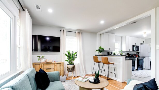 A modern living room with a sofa, a coffee table, and a TV. There is a kitchen in the background with a dining area.