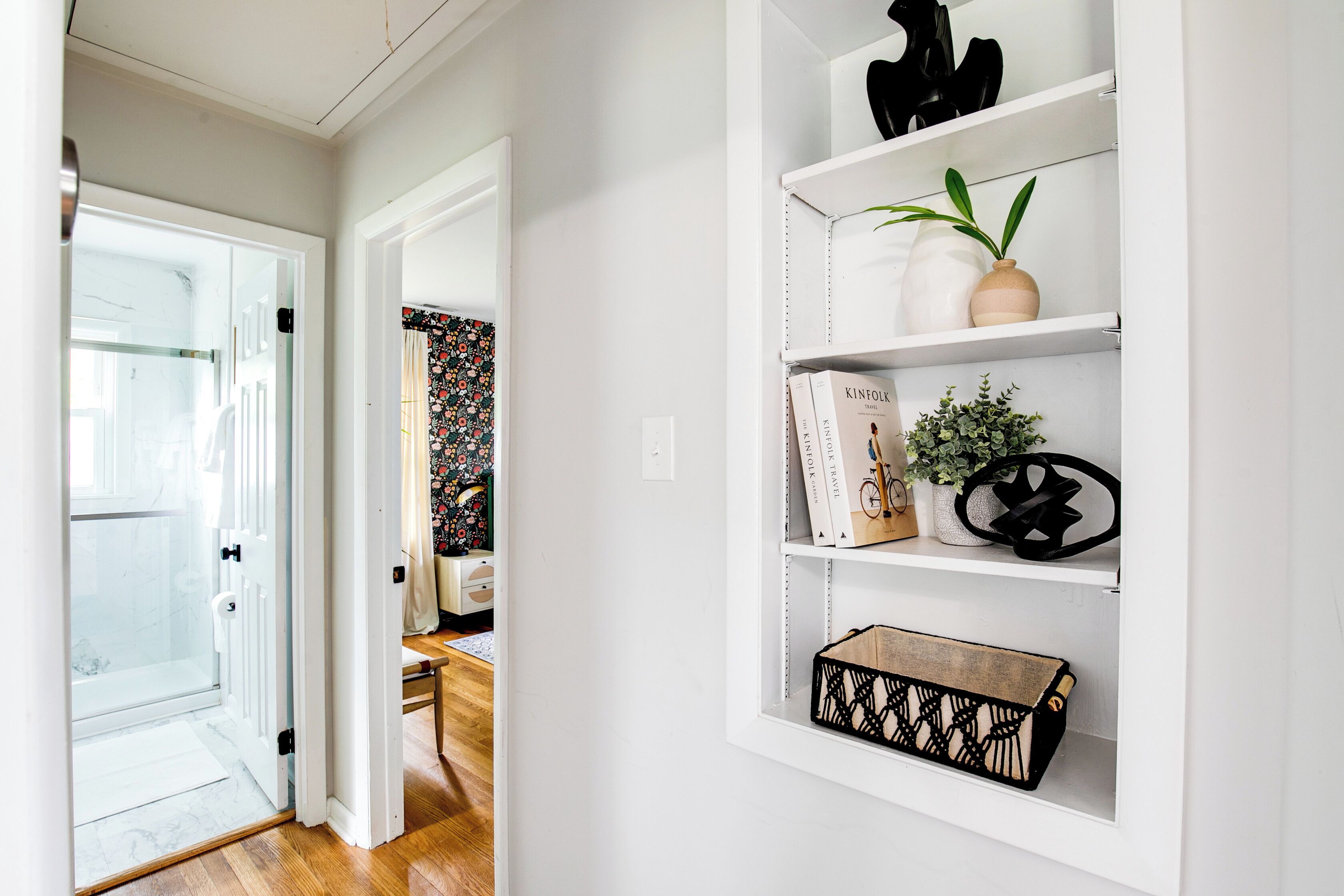 A hallway with a glass door leading to a bathroom, a white wall with a shelf holding books, a potted plant, and decorative items.