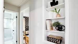A hallway with a glass door leading to a bathroom, a white wall with a shelf holding books, a potted plant, and decorative items.