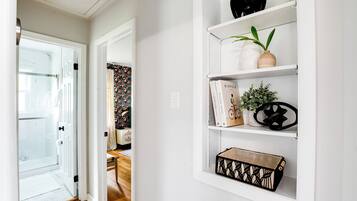 A hallway with a glass door leading to a bathroom, a white wall with a shelf holding books, a potted plant, and decorative items.