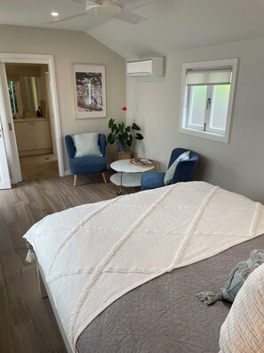 A bedroom with a large bed, two chairs, a small table, a potted plant, and a window with blinds. - Bryon's secret hideout studio (Byron Bay)