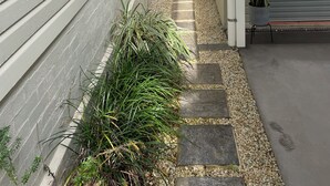 Un étroit sentier bordé d’une maison blanche avec une porte de garage, d’un petit banc en bois et d’une plante en pot.