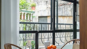 Varanda com cadeiras de vime, uma mesa redonda pequena com um bule e laranjas, e vista para um prédio com plantas na fachada.