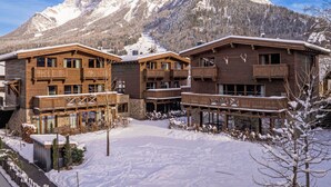 Une cour enneigée, avec des bâtiments en bois et une chaîne de montagnes en arrière-plan.