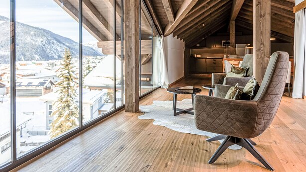 Ein geräumiger Raum mit hoher Decke und Holzbalken, ausgestattet mit einem großen Fenster mit Blick auf eine verschneite Landschaft und einer gemütlichen Sitzecke mit einem modernen Sessel und einem kleinen Tisch.