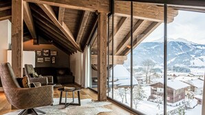 A spacious room with wooden beams, a leather armchair, a small table, and a large window offering a view of a snowy mountain landscape.